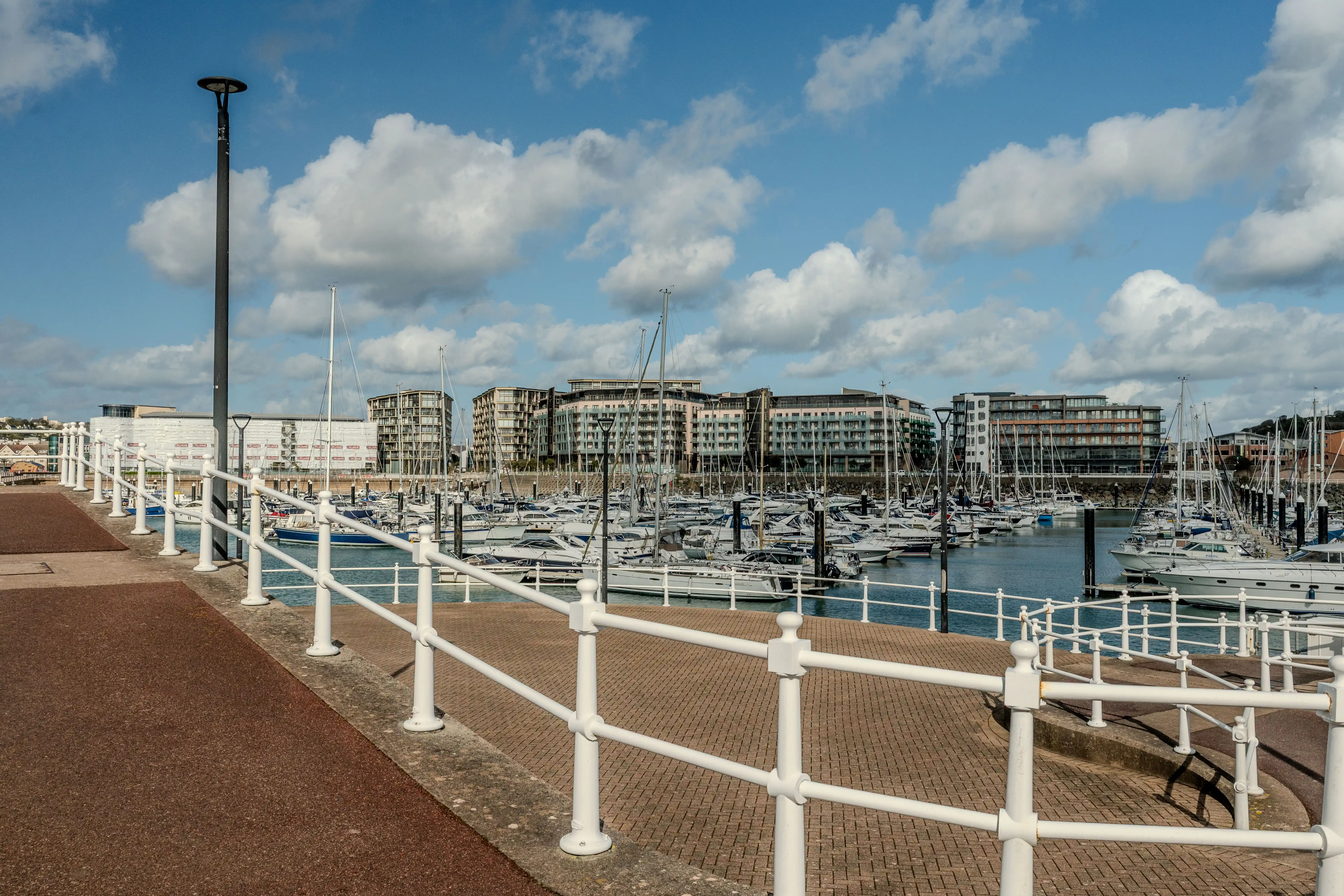 A marina filled with boats, with modern apartment buildings in the background and a pathway with a white railing in the foreground under a partly cloudy sky.